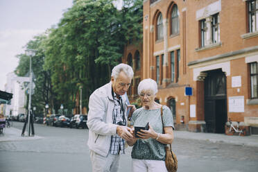 Senior woman holding smart phone discussing with partner while exploring in city - MASF17684