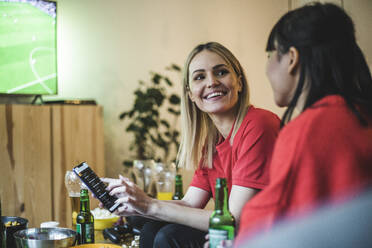 Smiling woman with smart phone looking at friend while watching soccer at home - MASF17818