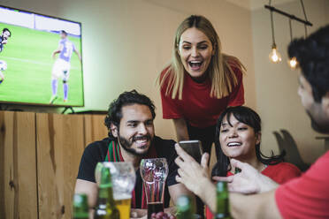 Smiling friends looking at smart phone while watching soccer in living room - MASF17821