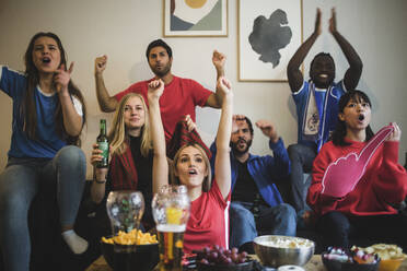 Happy friends with arms raised watching sport while sitting on sofa at home - MASF17837