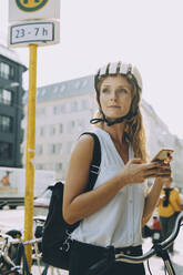 Junge Frau mit Fahrrad, die wegschaut und ihr Smartphone in der Stadt hält - MASF17839