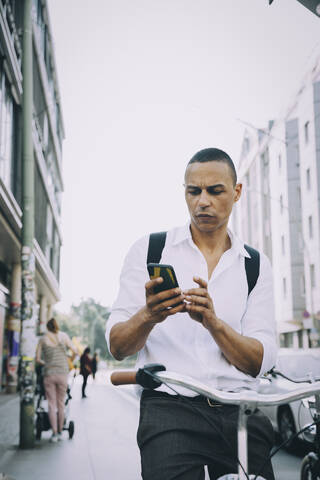 Geschäftsmann, der ein Mobiltelefon benutzt, während er mit dem Fahrrad auf dem Bürgersteig in der Stadt fährt, lizenzfreies Stockfoto