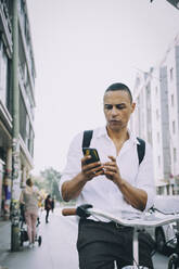 Geschäftsmann, der ein Mobiltelefon benutzt, während er mit dem Fahrrad auf dem Bürgersteig in der Stadt fährt - MASF17847