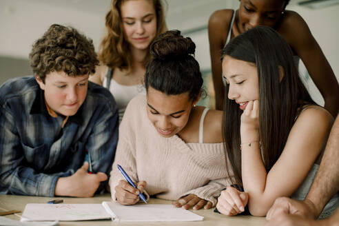 Lächelndes Teenager-Mädchen schreibt in ein Buch, während es inmitten von Freunden im Klassenzimmer sitzt - MASF17880