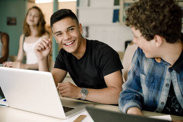 Lächelnde männliche Studenten, die einen Laptop über dem Tisch benutzen, während sie im Klassenzimmer sitzen - MASF17889