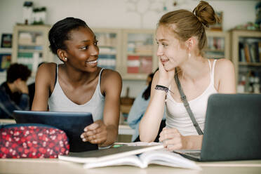 Smiling teenagers talking while sitting by table in classroom - MASF17892