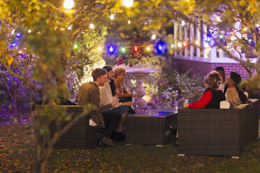 Freunde unterhalten sich und trinken unter Bäumen mit Lichterketten bei einer Gartenparty - CAIF25993