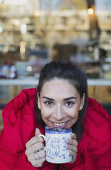 Porträt einer lächelnden, selbstbewussten jungen Frau, die im Fenster eines Cafés Kaffee trinkt - CAIF26060