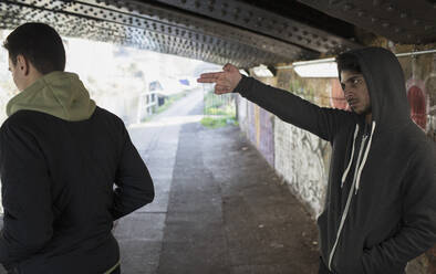 Menacing young man gesturing finger gun at man in urban tunnel - CAIF26155