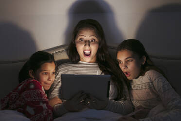 Überrascht Mutter und Töchter beobachten Film auf digitalen Tablet in dunklen Schlafzimmer - CAIF26218
