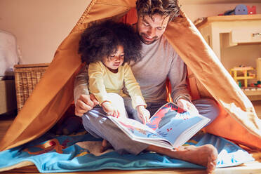 Father and daughter reading book in teepee - CAIF26242