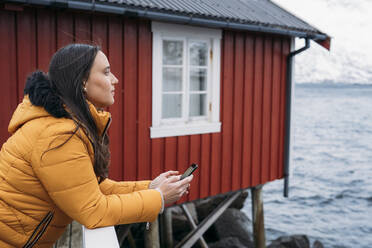 Tourist mit Mobiltelefon an einer Hütte an der Küste, Lofoten, Norwegen - MPPF00709