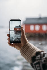 Nahaufnahme eines Touristen, der mit seinem Handy ein Foto von einer Hütte an der Küste macht, Lofoten, Norwegen - MPPF00729