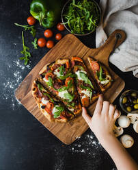 Draufsicht auf eine Hand, die nach einem Stück handgemachter Pizza auf einem Holzbrett greift. - CAVF78540