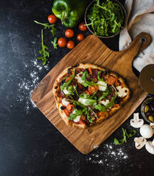 Draufsicht auf eine handgemachte Pizza auf einem Holzbrett mit Zutaten drum herum. - CAVF78541