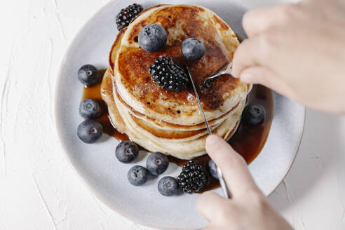 Frau schneidet eine Scheibe von einem Stapel Pfannkuchen mit Beeren und Sirup ab - CAVF78565