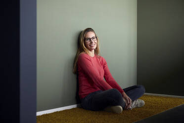 Portrait of smiling woman with glasses sitting on the floor - RBF07503