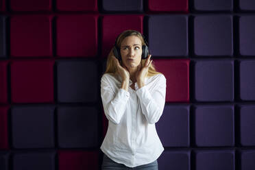 Porträt einer Frau an einer lila Wand, die mit Kopfhörern Musik hört - RBF07542