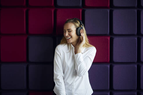 Porträt einer glücklichen Frau an einer lila Wand, die mit Kopfhörern Musik hört - RBF07544