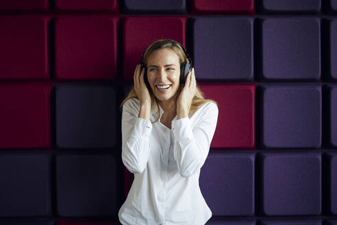 Porträt einer glücklichen Frau an einer lila Wand, die mit Kopfhörern Musik hört - RBF07545