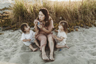 Mother with young twin daughters blowing bubbles at the beach - CAVF78943