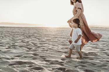 Side view of mother walking toddler twins to ocean with sun setting - CAVF78948