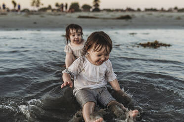 Front view of toddler sisters sitting and splashing in water at beach - CAVF78949