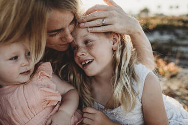 Portrait of mother cuddling with young daughters at beach sunset - CAVF78962