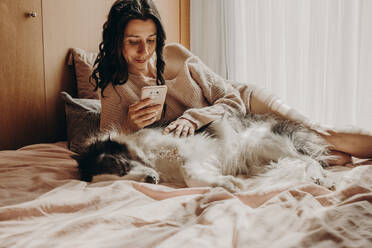 Porträt einer Frau, die mit ihrem Hund auf dem Bett liegt und auf ihr Smartphone schaut - GMLF00002