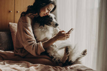 Lächelnde Frau sitzt mit ihrem Hund auf dem Bett und macht ein Selfie mit ihrem Smartphone - GMLF00005