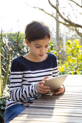 Portrait of little girl sitting at garden table looking at digital tablet - LVF08767
