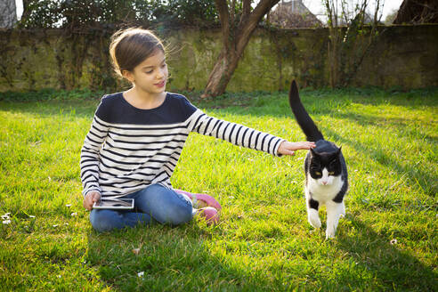 Porträt eines kleinen Mädchens, das mit einem Mini-Tablet auf einer Wiese sitzt und eine Katze streichelt - LVF08770