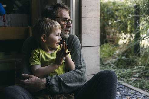 Vater und kleiner Sohn sitzen zu Hause auf dem Boden und schauen aus dem Fenster - MFF05407