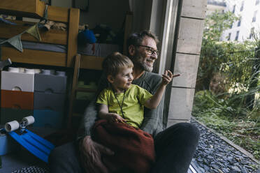 Father and little son sitting on the floor at home looking out of window - MFF05408