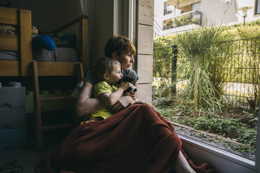 Mother and little son sitting on the floor at home looking out of window - MFF05411