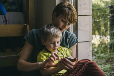 Mutter und kleiner Sohn kuscheln zu Hause und schauen auf ihr Smartphone - MFF05413