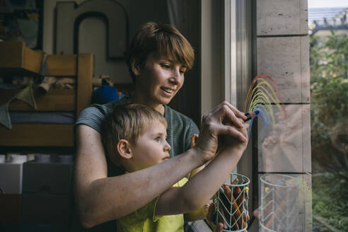 Mother and little son drawing rainbow on window at home - MFF05414
