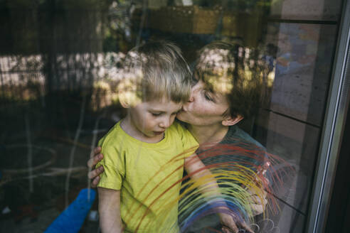 Mutter küsst ihren kleinen Sohn hinter dem Fenster mit gezeichnetem Regenbogen - MFF05416