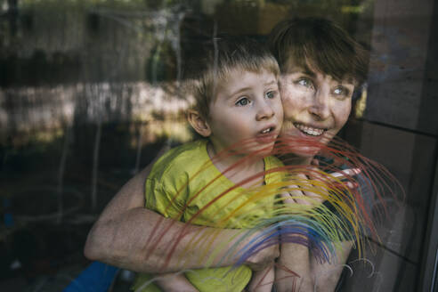 Porträt einer glücklichen Mutter und ihres kleinen Sohnes, die aus dem Fenster mit gezeichnetem Regenbogen schauen - MFF05418