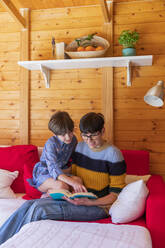 Mother and daughter reading a book on couch in a wooden cabin - VSMF00033