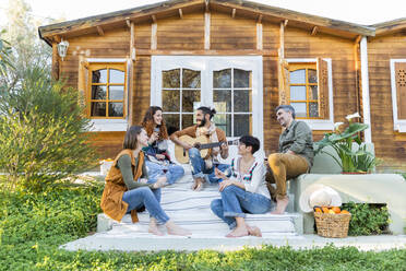 Friends playing music on the guitar and drinking wine outside a cabin in the countryside - VSMF00049