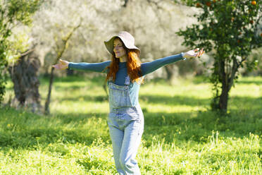 Redheaded woman wearing a hat enjoying the countryside with open arms - VSMF00139