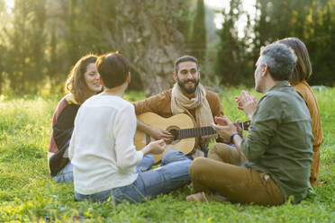 Gruppe von Freunden, die im Gras sitzend auf einem Feld mit der Gitarre Musik machen - VSMF00167