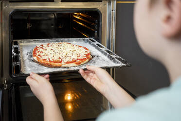 Ausschnittansicht eines Jungen, der ein Backblech mit roher Pizza in den Ofen schiebt - VPIF02225