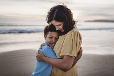Portrait of beautiful mother hugging young son at beach during sunset - CAVF79000