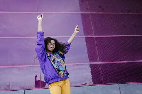 Young woman with urban look cheering in front of pink glass wall - TCEF00465