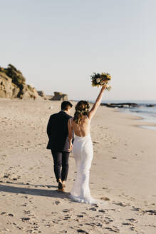 Rückansicht eines glücklichen Brautpaares beim Spaziergang am Strand - LHPF01254