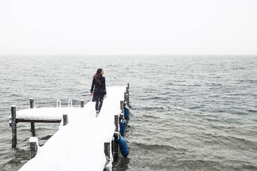 Rückenansicht einer jungen Frau, die auf einem schneebedeckten Steg am Starnberger See spazieren geht, Deutschland - WFF00324