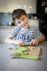 Porträt eines lächelnden kleinen Jungen, der in der Küche grünen Spargel schneidet - HMEF00868