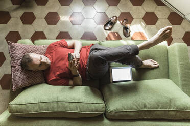 Mature man lying on sofa in living room and using tablet - MCVF00273